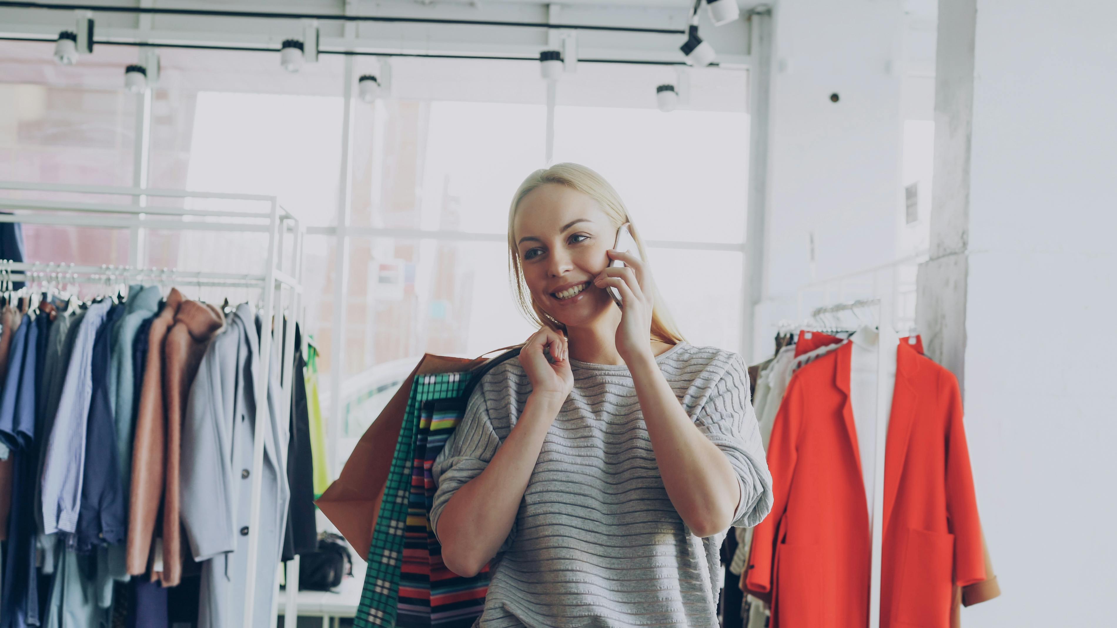 Smiling woman talks on phone while shopping in a bright clothing store.